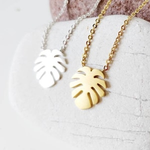 Puede incluir: Dos delicados collares con un colgante de hoja tropical. Un collar es dorado y el otro es plateado.