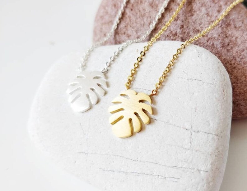 Puede incluir: Dos delicados collares con un colgante de hoja tropical. Un collar es plateado y el otro es dorado.