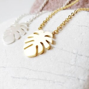 Puede incluir: Dos collares de oro y plata con un colgante de hoja de monstera. Los collares est&aacute;n sobre una superficie de piedra blanca.