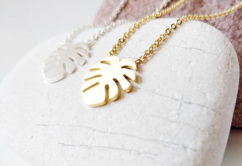 Puede incluir: Dos collares de oro y plata con un colgante de hoja de monstera. Los collares est&aacute;n sobre una superficie de piedra blanca.