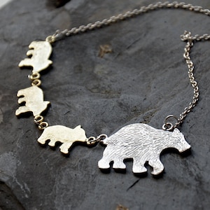May include: A necklace featuring a silver-toned chain connected to a series of four bear-shaped charms. The largest bear charm is textured and silver-colored, while the three smaller bear charms are smooth and gold-toned, linked together in a row.