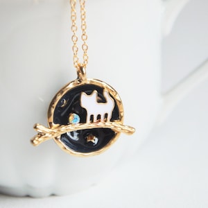May include: A gold necklace with a black and white enamel pendant featuring a cat walking on a branch. The pendant has a small, clear rhinestone.