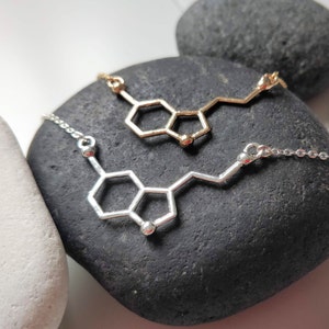 May include: Two necklaces with a silver and gold pendant in the shape of a dopamine molecule. The necklaces are displayed on two grey stones.