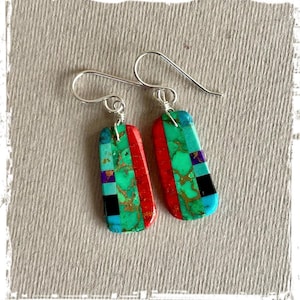 May include: A pair of rectangular earrings with a mosaic design. The earrings feature turquoise, red, black, purple, and gold-colored stones. The earrings have silver-colored ear wires.