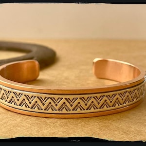 May include: Copper cuff bracelet with a geometric design. The bracelet has a polished copper finish with a band of gold-colored metal featuring a repeating black zigzag pattern. The open cuff design allows for easy wear.