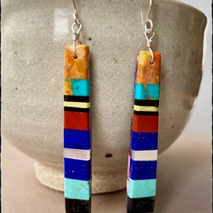 May include: A pair of long, rectangular dangle earrings with a multi-colored inlay design. The earrings are made of silver and feature a variety of stones, including turquoise, lapis lazuli, and red jasper.