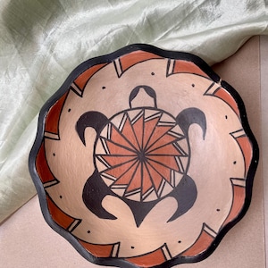 May include: A ceramic bowl with a black rim and a scalloped edge. The bowl is decorated with a stylized turtle design in black, orange, and white.