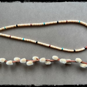 May include: A white shell necklace with red, black, and turquoise accents. The necklace is made of small, round beads and has a silver clasp.