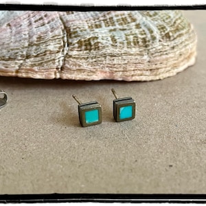May include: A pair of small square stud earrings featuring a bright turquoise inlay set within a dark metallic frame. The earrings include two separate metal earring backs, each with a circular design, positioned to the left of the studs.