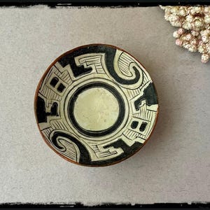 May include: A small, round ceramic bowl with a cream-colored interior and a black and white geometric design. The rim is a dark brown. The bowl's design features a central circle and repeating shapes. The bowl is likely handmade.