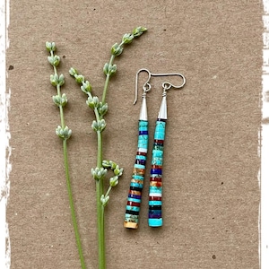 May include: A pair of handcrafted earrings with a silver hook and a cylindrical design. The earrings are made of stacked, colorful stones including turquoise, red, and black. A sprig of lavender is next to the earrings on a brown background.