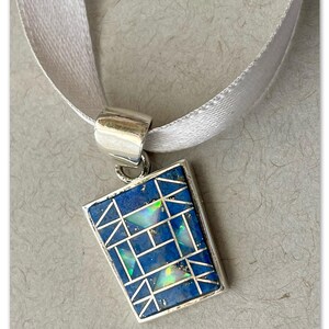 May include: A silver pendant with a geometric design featuring blue lapis lazuli and iridescent opal stones. The pendant is attached to a white satin ribbon.