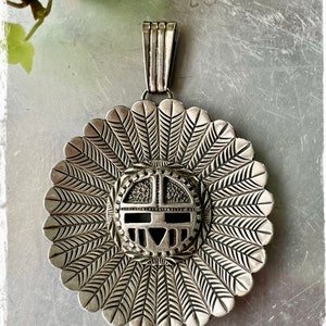 May include: A silver pendant with a detailed design of a Native American sun face with feathers around the edge.