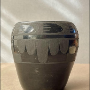 May include: Black pottery vase with geometric designs. The vase has a rounded body and a wide mouth.