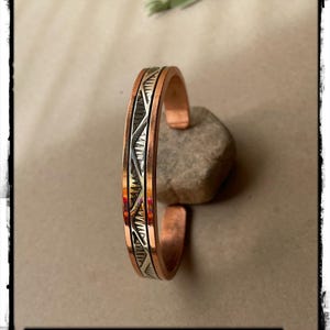 May include: A copper cuff bracelet featuring a central silver-toned inlay with a repeating geometric pattern of triangles and lines. The bracelet has a smooth, polished finish on the edges and a distinct open-ended design, showcasing a metallic contrast.