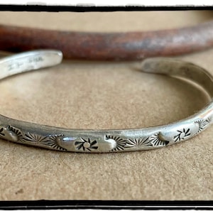 May include: A silver cuff bracelet featuring a series of stamped sunburst motifs and crescent shapes along the outer band. The interior of the metal cuff displays a small, stamped hallmark near the opening, showcasing a polished metallic finish.