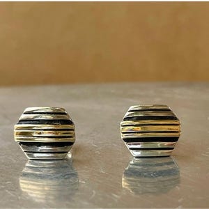 May include: A pair of stud earrings with a geometric design. Each earring features alternating bands of silver, black, and gold. The earrings are displayed on a reflective surface with a neutral background.