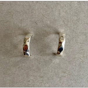 May include: Two silver-colored hoop earrings with a hammered texture. The earrings have a curved shape and a shiny finish, reflecting light. The earrings are displayed on a textured, neutral-toned surface.