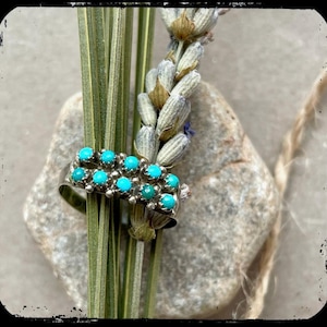 May include: A silver ring with two rows of turquoise stones. The ring is set against a backdrop of lavender and greenery, with a neutral stone in the background. The ring is a piece of jewelry.