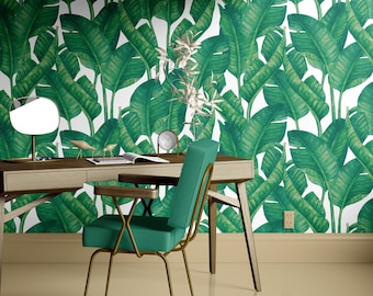 May include: A teal green chair with a gold metal frame sits in front of a wooden desk with a drawer. The desk is against a wall with a white background and a green banana leaf pattern wallpaper.