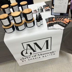 May include: A white table covered with a white tablecloth featuring a black and white logo that reads "Artz of Massage by Shakia Batten". The table is decorated with black candles with wooden lids, small bottles of massage oil, and brochures that read "Artz of Massage by Shakia Batten".