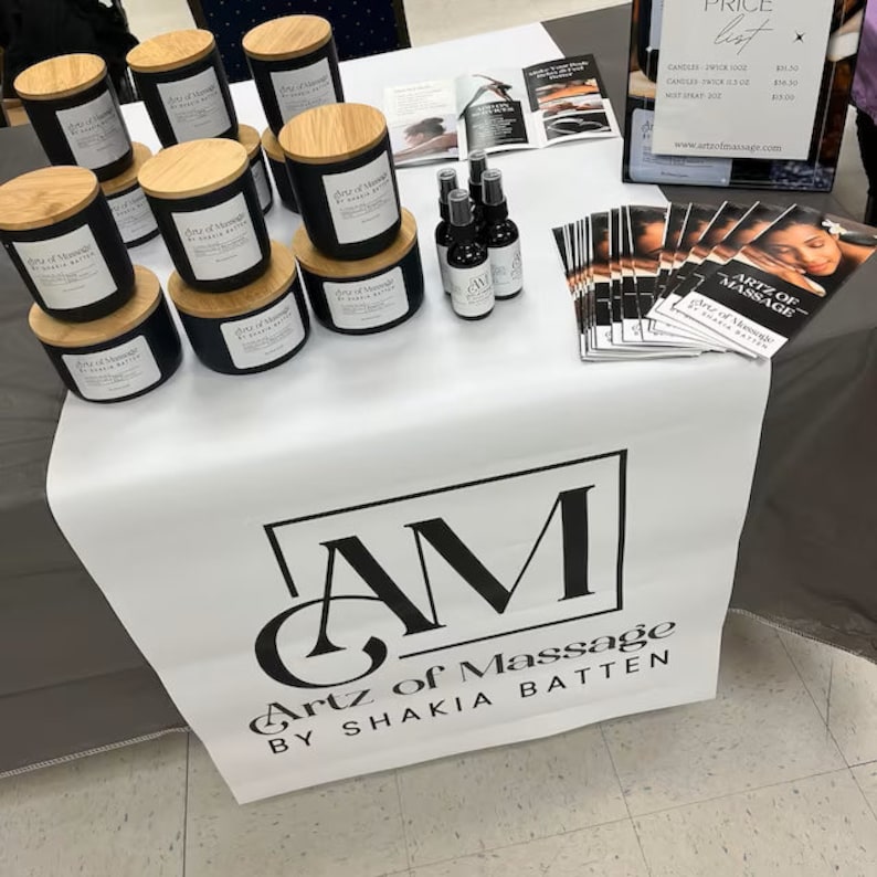 May include: A white table covered with a white tablecloth featuring a black and white logo that reads "Artz of Massage by Shakia Batten". The table is decorated with black candles with wooden lids, small bottles of massage oil, and brochures that read "Artz of Massage by Shakia Batten".