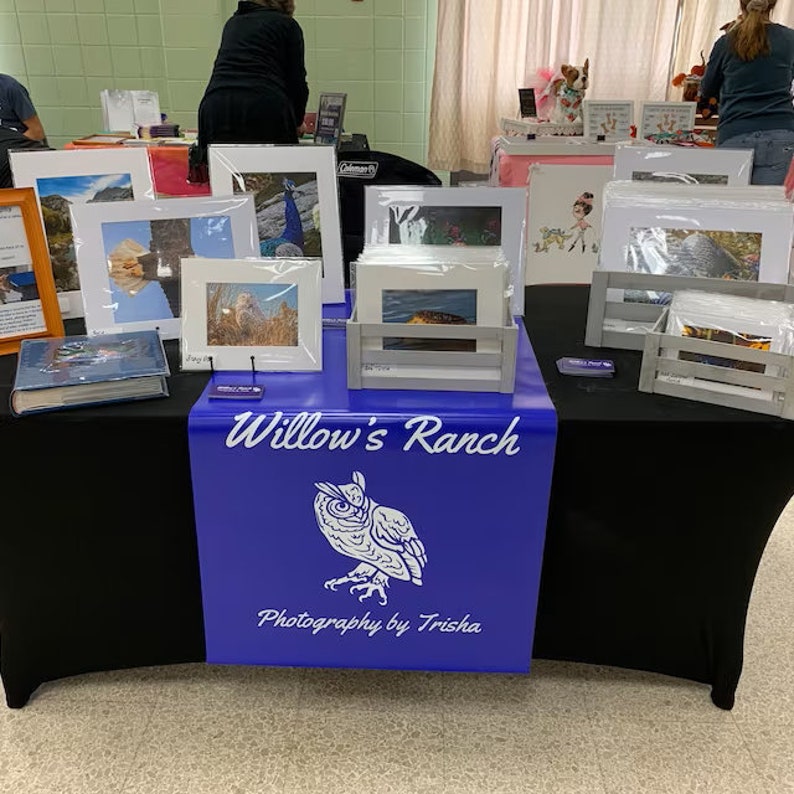 May include: A table covered in a black tablecloth with a blue table runner that says "Willow's Ranch Photography by Trisha". The table is decorated with framed photographs of nature scenes, including a peacock, a bird, and a landscape.