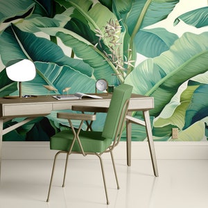 May include: A green desk chair with a metal frame and a wooden desk with a white top and a lamp with a white shade. The desk and chair are in front of a wall with a tropical banana leaf wallpaper.