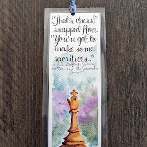 May include: A watercolor painting of a chess queen on a purple and green background. The quote "That's chess!" snapped Ron. "You've got to make some sacrifices." - J.K. Rowling, "Harry Potter and the Sorcerer's Stone" is written on the bookmark.