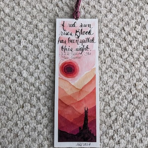 May include: A watercolor bookmark with a red sun rising over a mountain range. The quote "A red sun rises. Blood has been spilled this night." from J.R.R. Tolkien's "The Two Towers" is written in black ink. The bookmark is signed "M 2024".