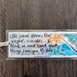 May include: A watercolor bookmark with a quote from the Harry Potter book "Harry Potter and the Sorcerer's Stone" by J.K. Rowling. The quote reads: "The wand chooses the wizard, remember... I think we must expect great things from you, Mr. Potter." The bookmark features a watercolor painting of a wand on a blue and green background.