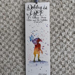 May include: A watercolour painting of Dobby the house-elf from the Harry Potter series. The painting is on a white background with a blue, yellow, orange, and red colour scheme. The text "Dobby is FREE" is written in black ink at the top of the bookmark. The text "J.K. Rowling, Harry Potter and the Chamber of Secrets" is written in black ink below the painting. The date "May 2024" is written in black ink at the bottom of the bookmark.