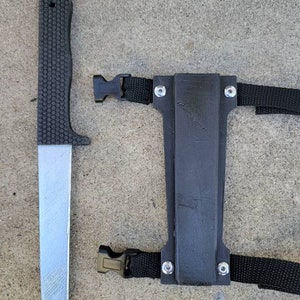 Puede incluir: Un cuchillo negro con mango texturizado y hoja plateada, junto a una funda negra con correas y hebillas ajustables. El cuchillo y la funda est&aacute;n sobre una superficie de hormig&oacute;n gris.