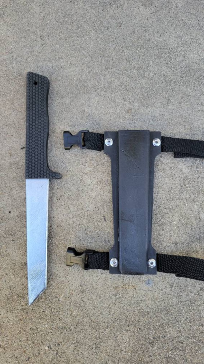Puede incluir: Un cuchillo negro con mango texturizado y hoja plateada, junto a una funda negra con correas y hebillas ajustables. El cuchillo y la funda est&aacute;n sobre una superficie de hormig&oacute;n gris.