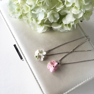 Peut inclure: Deux délicats colliers en argent avec des breloques en forme de fleurs. Un charme est blanc et l'autre est rose.