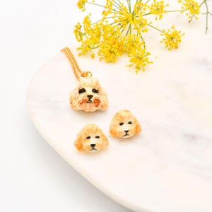May include: A gold-colored necklace with a pendant shaped like a dog's head, along with two matching dog head earrings. The dog's head is beige with black and brown details. The jewelry is displayed on a white marble surface with yellow flowers.