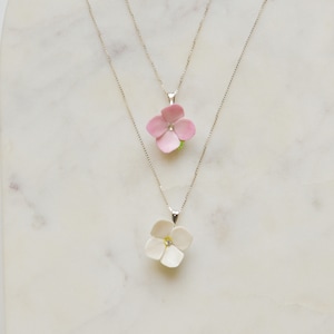 May include: Two silver necklaces with flower pendants. The top necklace has a pink flower pendant with a clear rhinestone in the centre. The bottom necklace has a white flower pendant with a clear rhinestone in the centre.