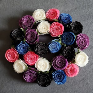 A photo of a coiled paper flower garland on a gray background. The garland is made up of small rolled roses in a repeating pattern of pink, white, purple, black, and blue, attached to a green leafy ribbon.