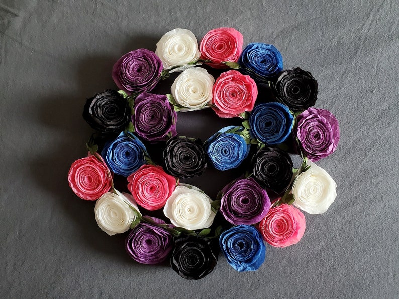 A photo of a coiled paper flower garland on a gray background. The garland is made up of small rolled roses in a repeating pattern of pink, white, purple, black, and blue, attached to a green leafy ribbon.