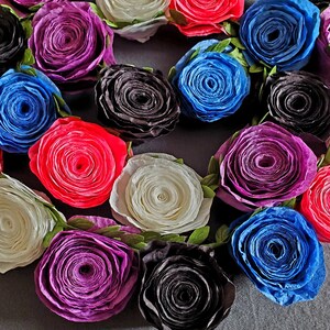 A photo of a coiled paper flower garland on a gray background. The garland is made up of small rolled roses in a repeating pattern of pink, white, purple, black, and blue, attached to a green leafy ribbon.