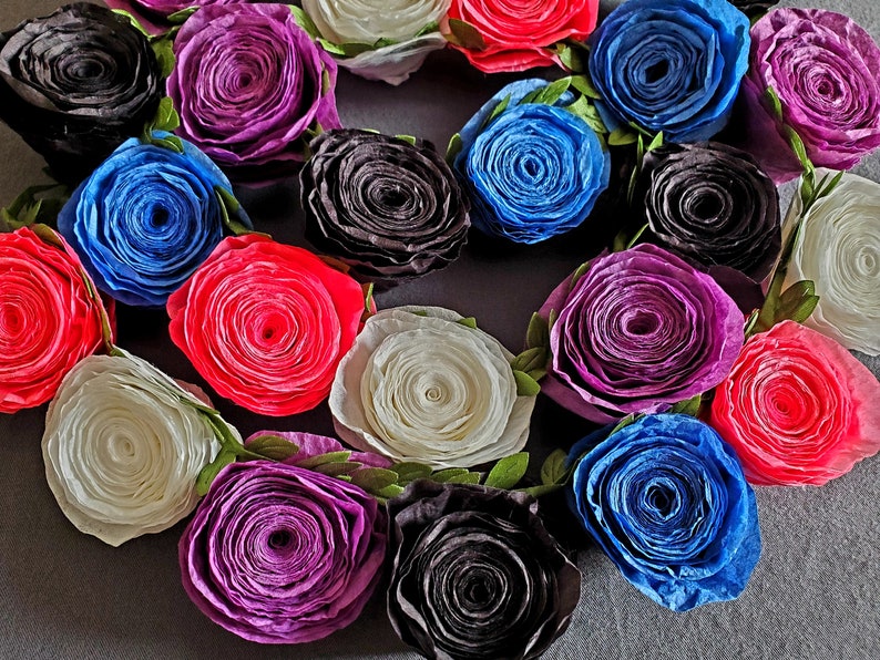 A photo of a coiled paper flower garland on a gray background. The garland is made up of small rolled roses in a repeating pattern of pink, white, purple, black, and blue, attached to a green leafy ribbon.