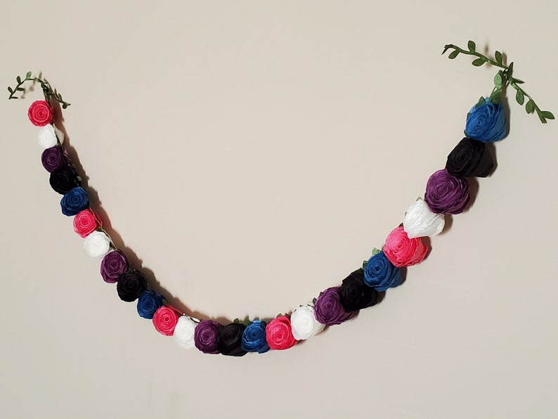 A photo of a paper flower garland hanging on a white wall. The garland is made up of small rolled roses in a repeating pattern of pink, white, purple, black, and blue, attached to a green leafy ribbon and is about 3 feet long.