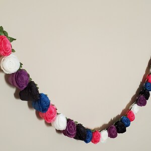 A photo of a paper flower garland hanging on a white wall. The garland is made up of small rolled roses in a repeating pattern of pink, white, purple, black, and blue, attached to a green leafy ribbon and is about 3 feet long.