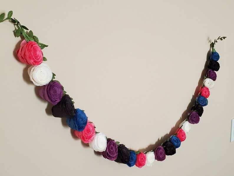 A photo of a paper flower garland hanging on a white wall. The garland is made up of small rolled roses in a repeating pattern of pink, white, purple, black, and blue, attached to a green leafy ribbon and is about 3 feet long.