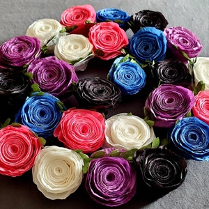 A photo of a coiled paper flower garland on a gray background. The garland is made up of small rolled roses in a repeating pattern of pink, white, purple, black, and blue, attached to a green leafy ribbon.