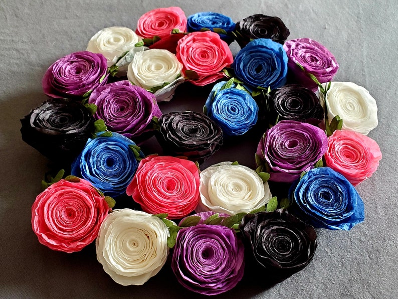 A photo of a coiled paper flower garland on a gray background. The garland is made up of small rolled roses in a repeating pattern of pink, white, purple, black, and blue, attached to a green leafy ribbon.