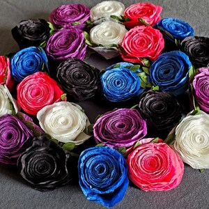 A photo of a coiled paper flower garland on a gray background. The garland is made up of small rolled roses in a repeating pattern of pink, white, purple, black, and blue, attached to a green leafy ribbon.