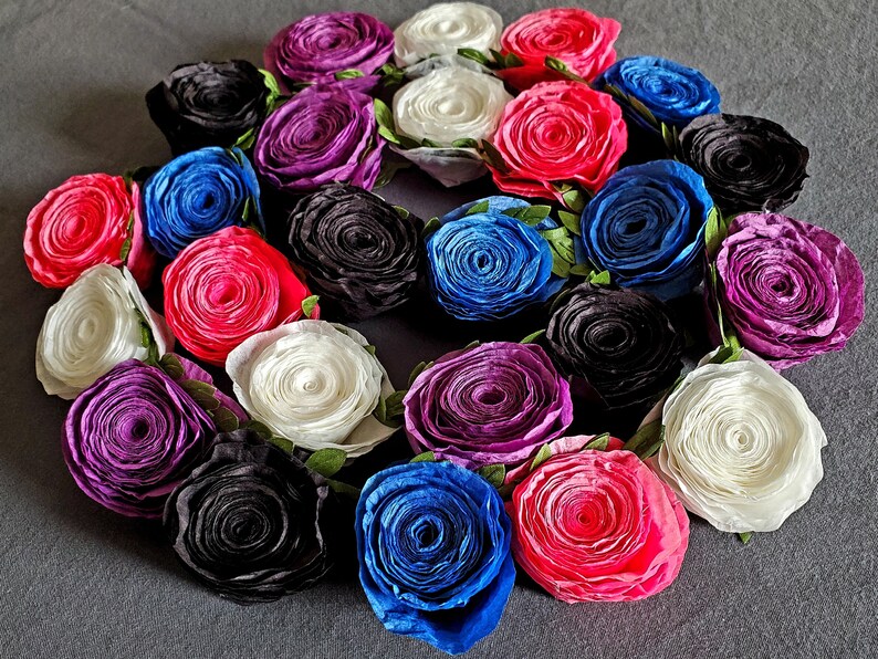 A photo of a coiled paper flower garland on a gray background. The garland is made up of small rolled roses in a repeating pattern of pink, white, purple, black, and blue, attached to a green leafy ribbon.