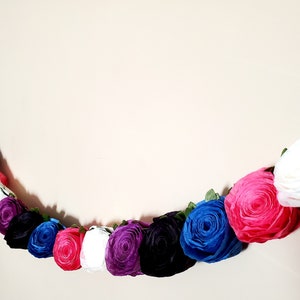 A photo of a paper flower garland hanging on a white wall. The garland is made up of small rolled roses in a repeating pattern of pink, white, purple, black, and blue, attached to a green leafy ribbon and is about 3 feet long.