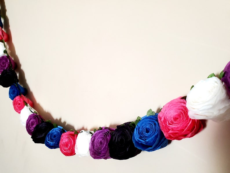 A photo of a paper flower garland hanging on a white wall. The garland is made up of small rolled roses in a repeating pattern of pink, white, purple, black, and blue, attached to a green leafy ribbon and is about 3 feet long.